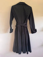 A black long-sleeve wrap dress with a matching waist tie belt, shown from the back hanging against a plain off-white wall.