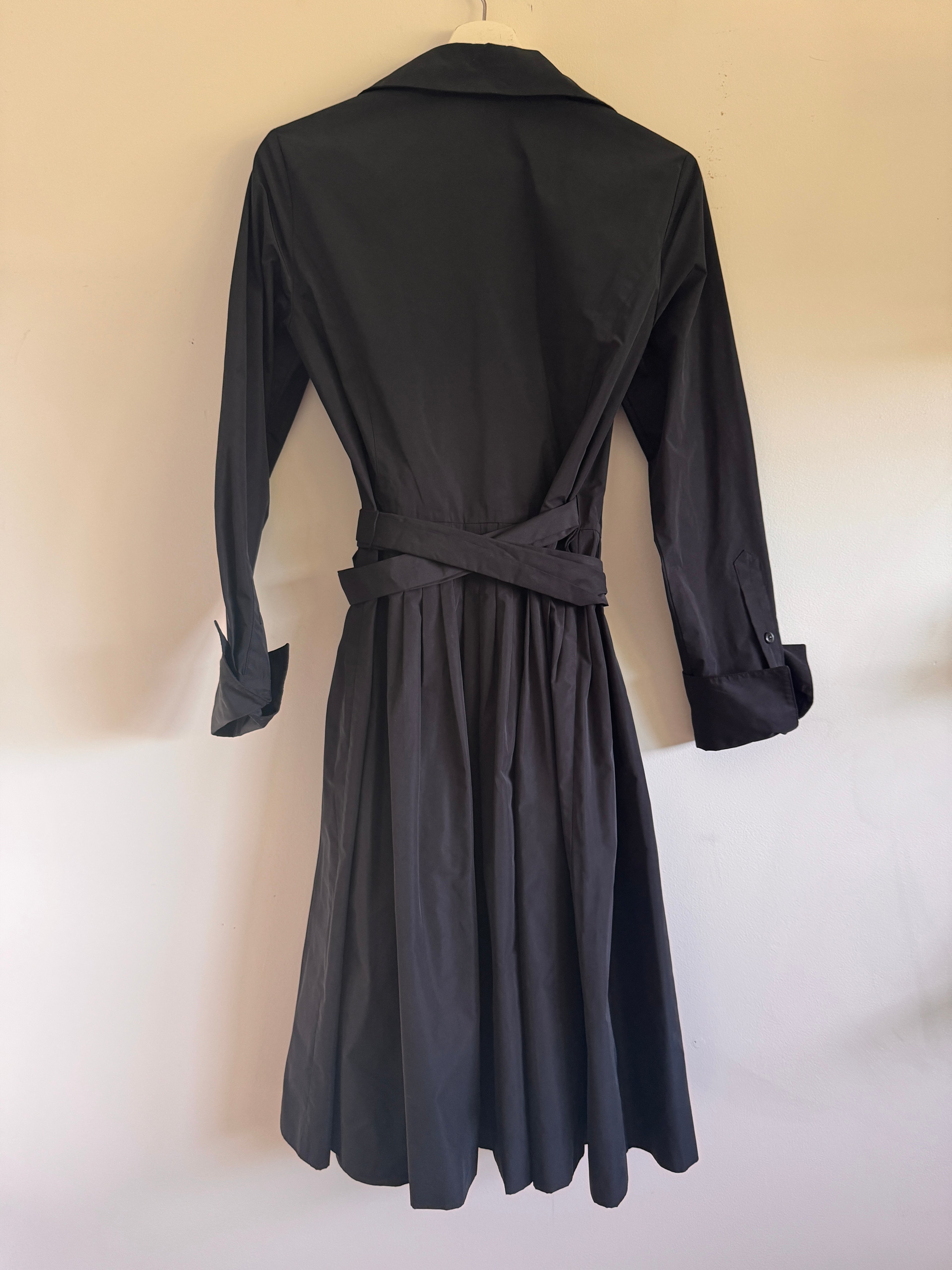 A black long-sleeve wrap dress with a matching waist tie belt, shown from the back hanging against a plain off-white wall.
