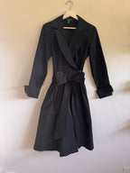 A black wrap-style dress with a self-tie belt and long sleeves, hanging on a wooden hanger against a plain off-white wall.