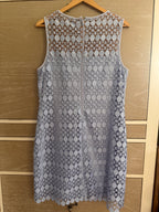 A sleeveless light blue lace dress with a circular pattern and a front zipper, hanging on a wooden hanger against a light wood paneled door.
