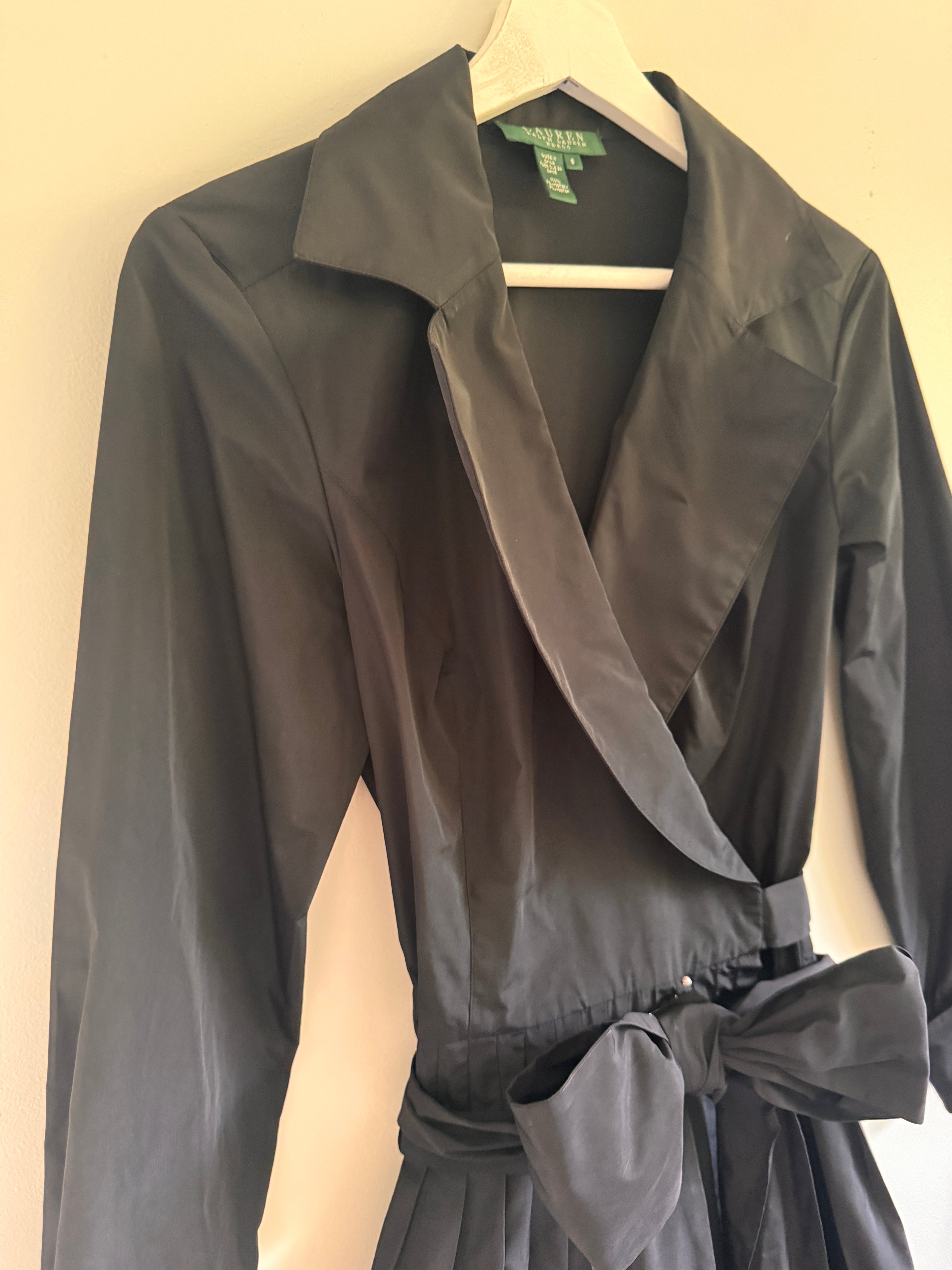Black wrap-style long-sleeve dress with a tie waist, hanging on a white hanger against a plain light beige wall.