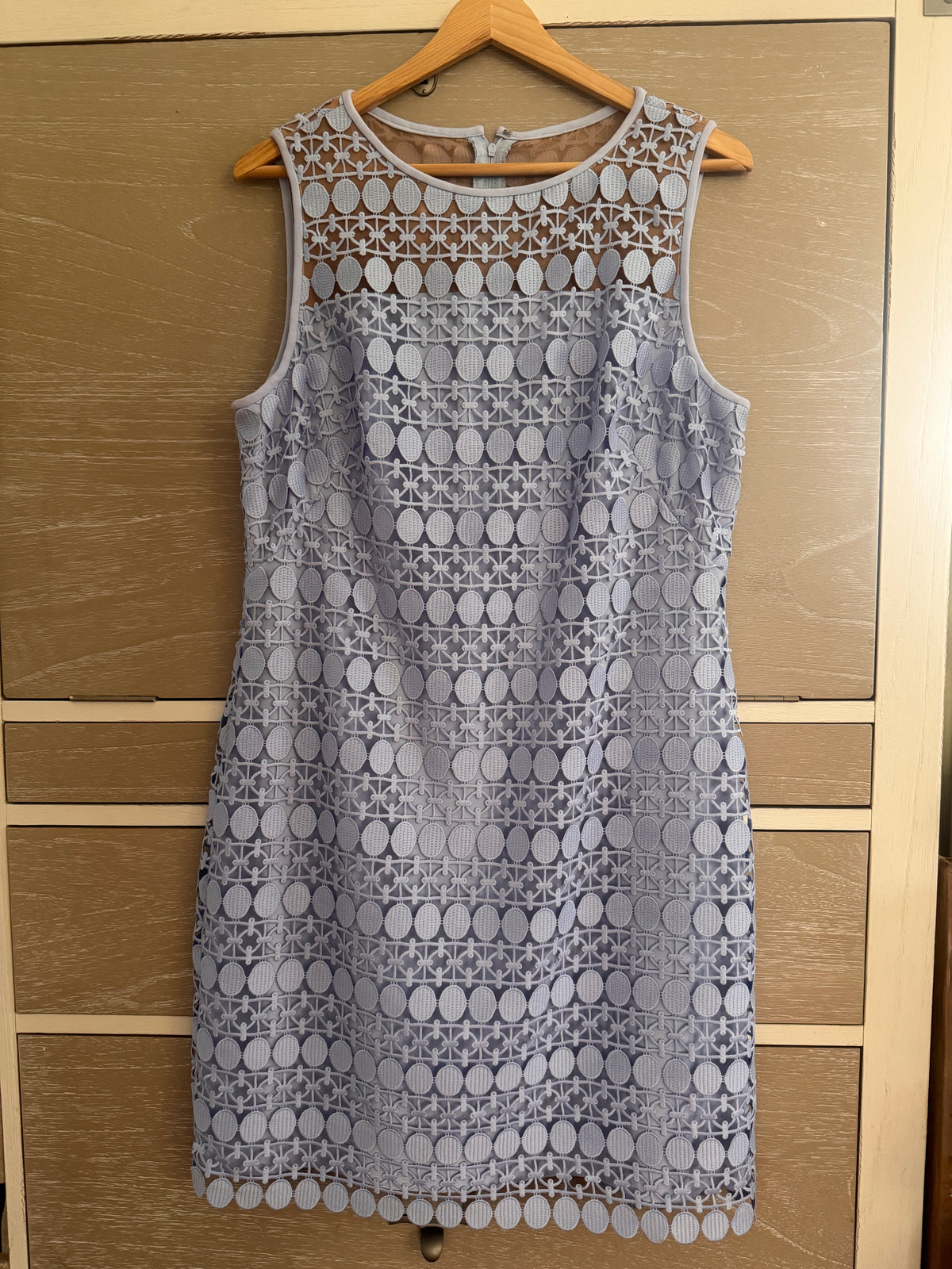 A sleeveless light blue lace dress with a scalloped hem and geometric floral lace pattern, hanging on a wooden hanger against a light wood drawer or cabinet background.