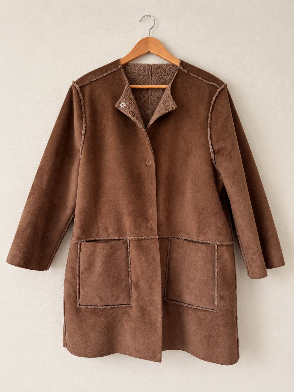 A brown suede-like jacket with a stand collar, button front, and two large front patch pockets, hanging on a wooden hanger against a plain light beige wall.