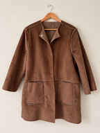 A brown suede-like jacket with a stand collar, button front, and two large front patch pockets, hanging on a wooden hanger against a plain light beige wall.