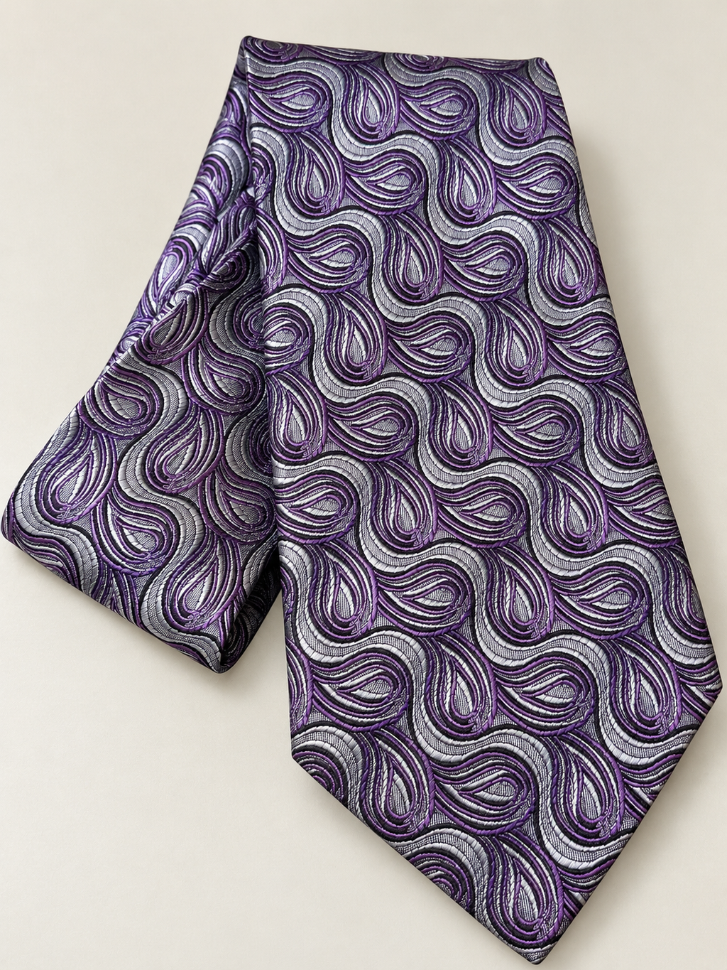 A purple and silver paisley patterned necktie, folded to show its texture and pattern, set against a plain light beige background.