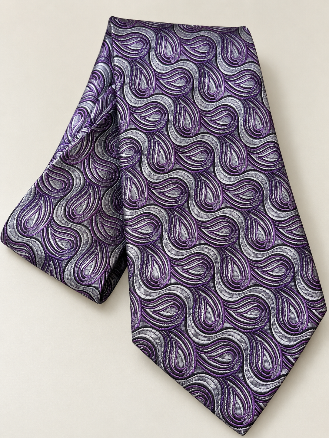 A purple and silver paisley patterned necktie, folded to show its texture and pattern, set against a plain light beige background.