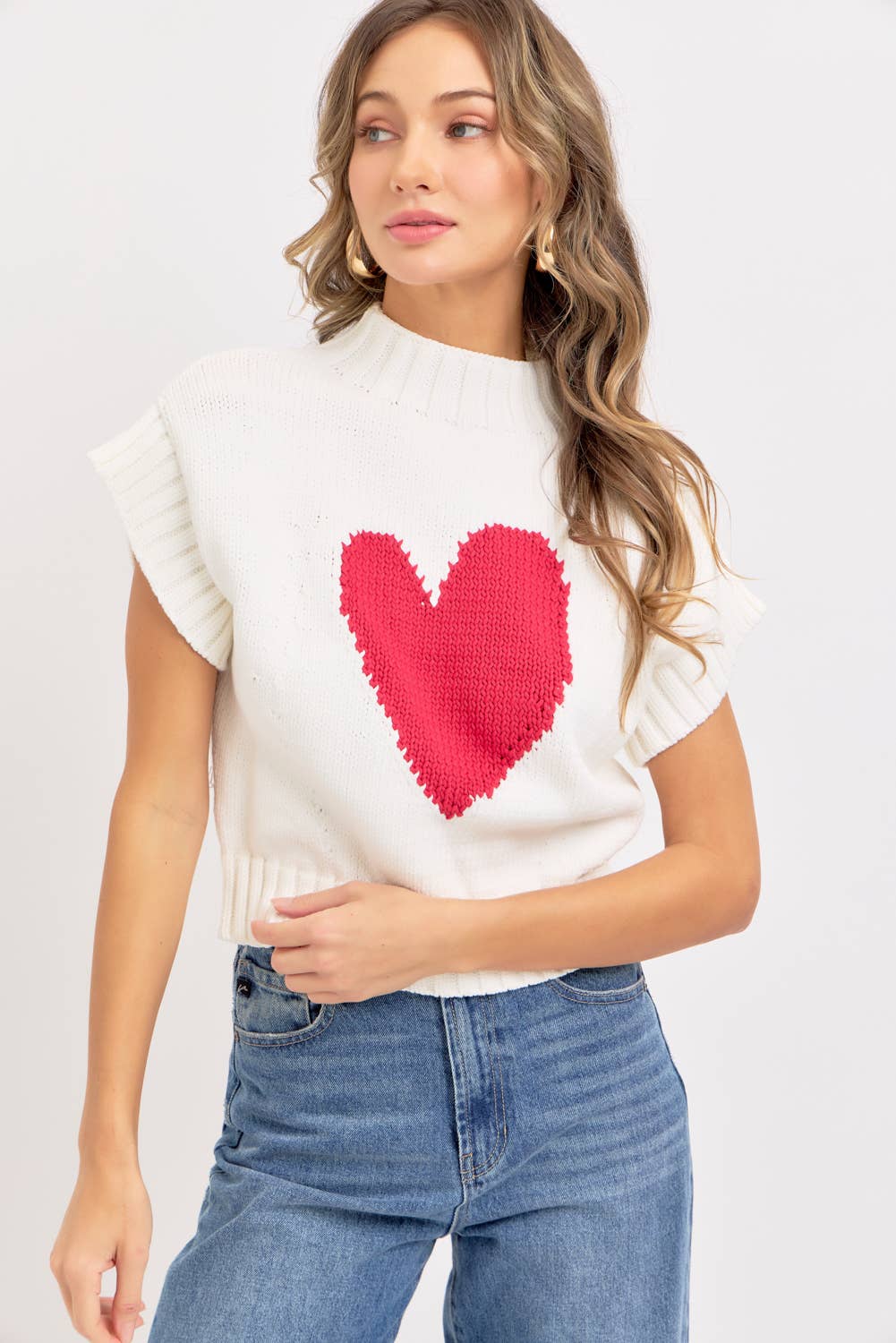 Woman wearing a white top with a red heart design and blue jeans on a plain background