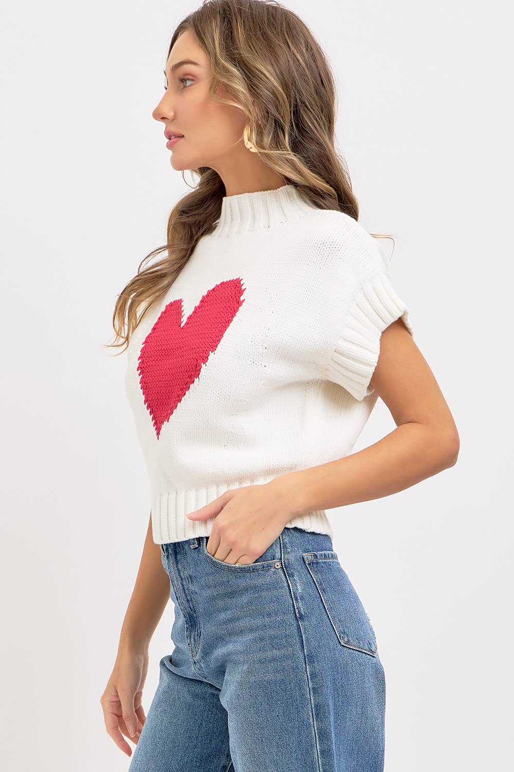 Woman wearing a white sleeveless sweater with a red heart design on a plain background