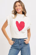 Woman wearing a white sleeveless sweater with a pink heart design on a plain background