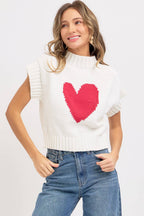 Woman wearing a white sleeveless sweater with a pink heart design on a plain background