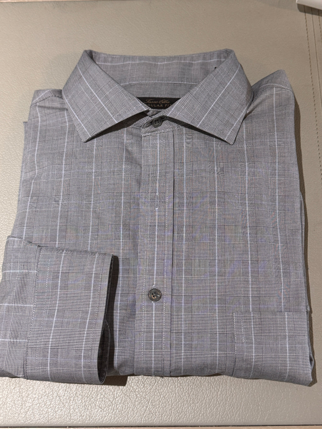 A folded gray long-sleeve dress shirt with a subtle white windowpane plaid pattern, displayed on a light beige textured surface.