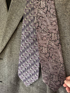Two patterned neckties displayed against a gray textured blazer. One tie features a wavy, abstract pattern in purple, gray, and white, while the other features a classic paisley pattern in dusty purple and black.