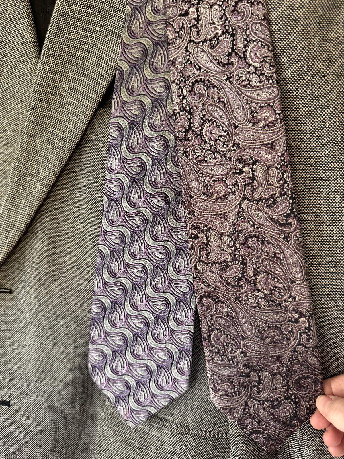 Two patterned neckties displayed against a gray textured blazer. One tie features a wavy, abstract pattern in purple, gray, and white, while the other features a classic paisley pattern in dusty purple and black.
