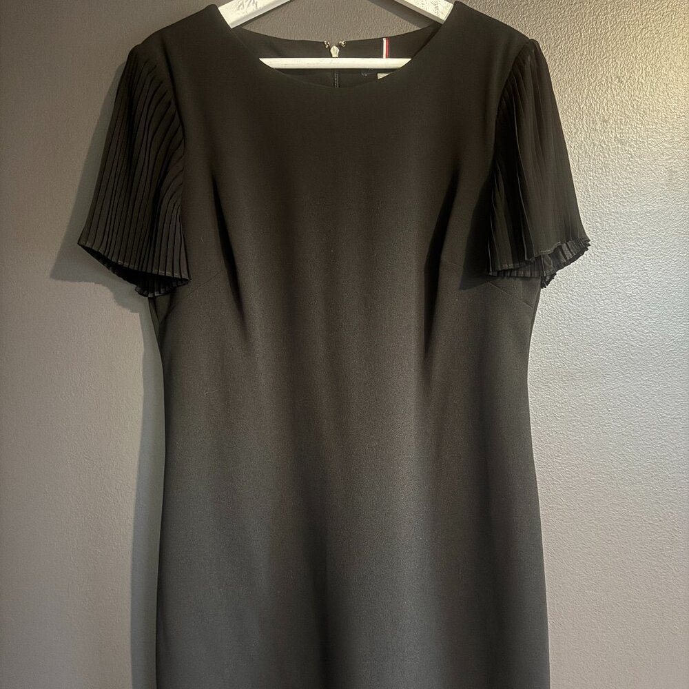 Black dress with pleated shoulders on a hanger against a neutral wall