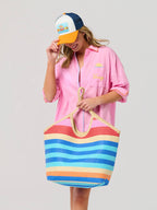 Woman wearing a pink dress and colorful cap, holding a striped beach bag on a plain background