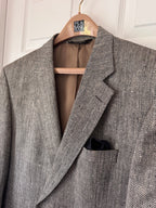 A black and brown textured herringbone blazer jacket hanging on a wooden hanger against a white paneled door, with a black pocket square in the chest pocket.