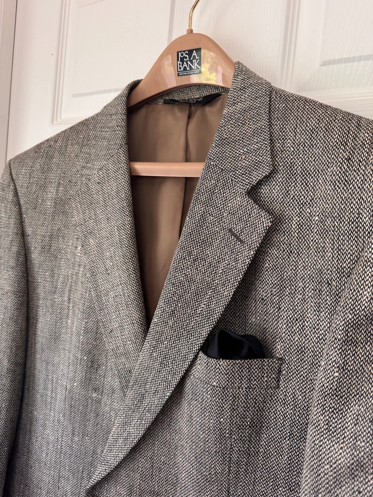 A black and brown textured herringbone blazer jacket hanging on a wooden hanger against a white paneled door, with a black pocket square in the chest pocket.