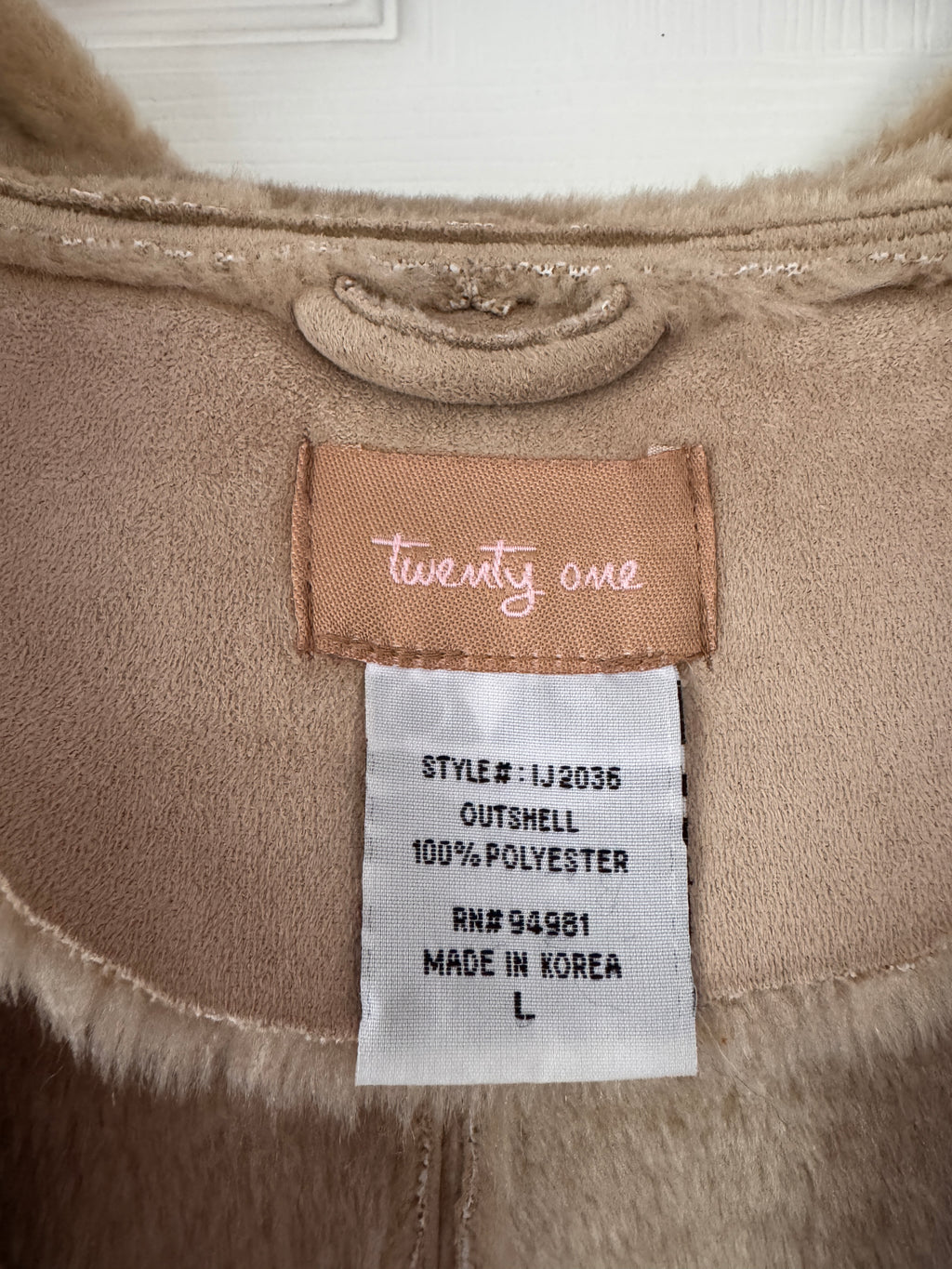 Close-up of the inner collar of a beige faux-fur jacket, showing a tan brand label reading 'twenty one' and a white size tag with product details.