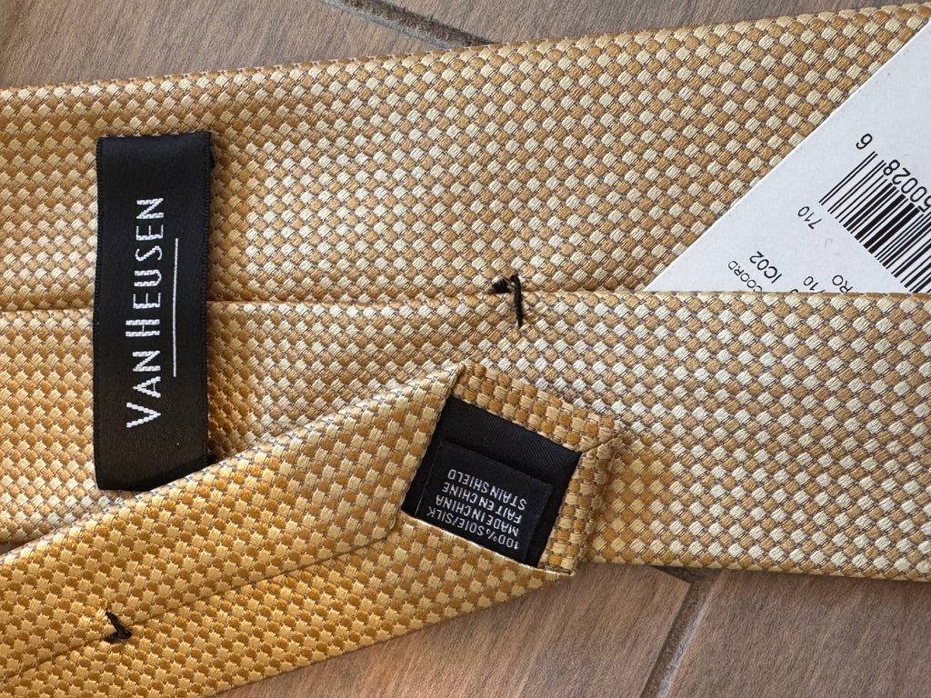 A close-up of a woven Van Heusen necktie in a textured beige and gold pattern, showing the black brand label and a white product tag on a wooden surface.