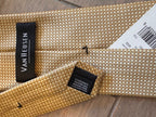 A close-up of a woven Van Heusen necktie in a textured beige and gold pattern, showing the black brand label and a white product tag on a wooden surface.