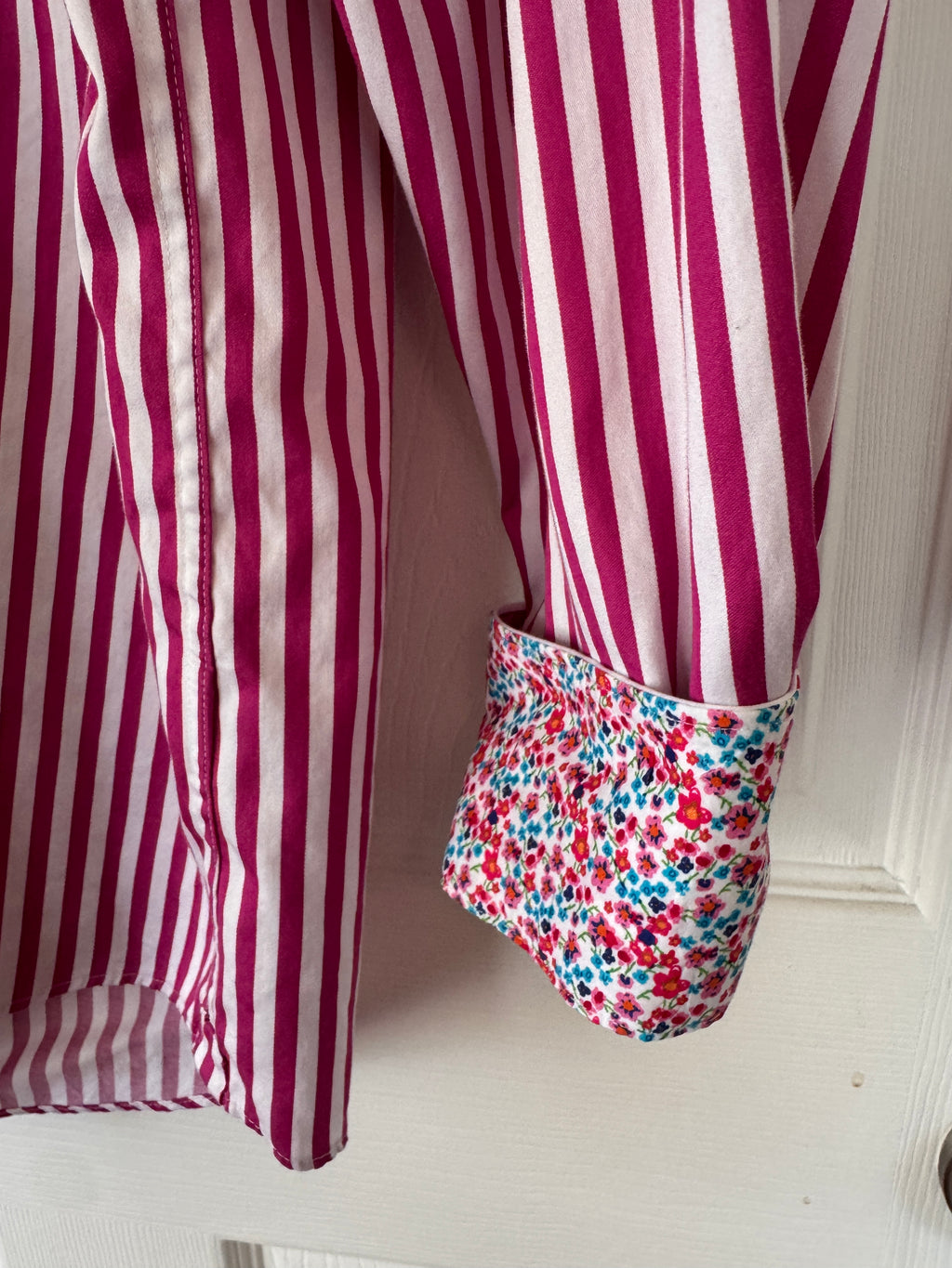 A close-up of a striped shirt with a rolled-up cuff showing a contrasting floral patterned lining, hanging against a white paneled door.