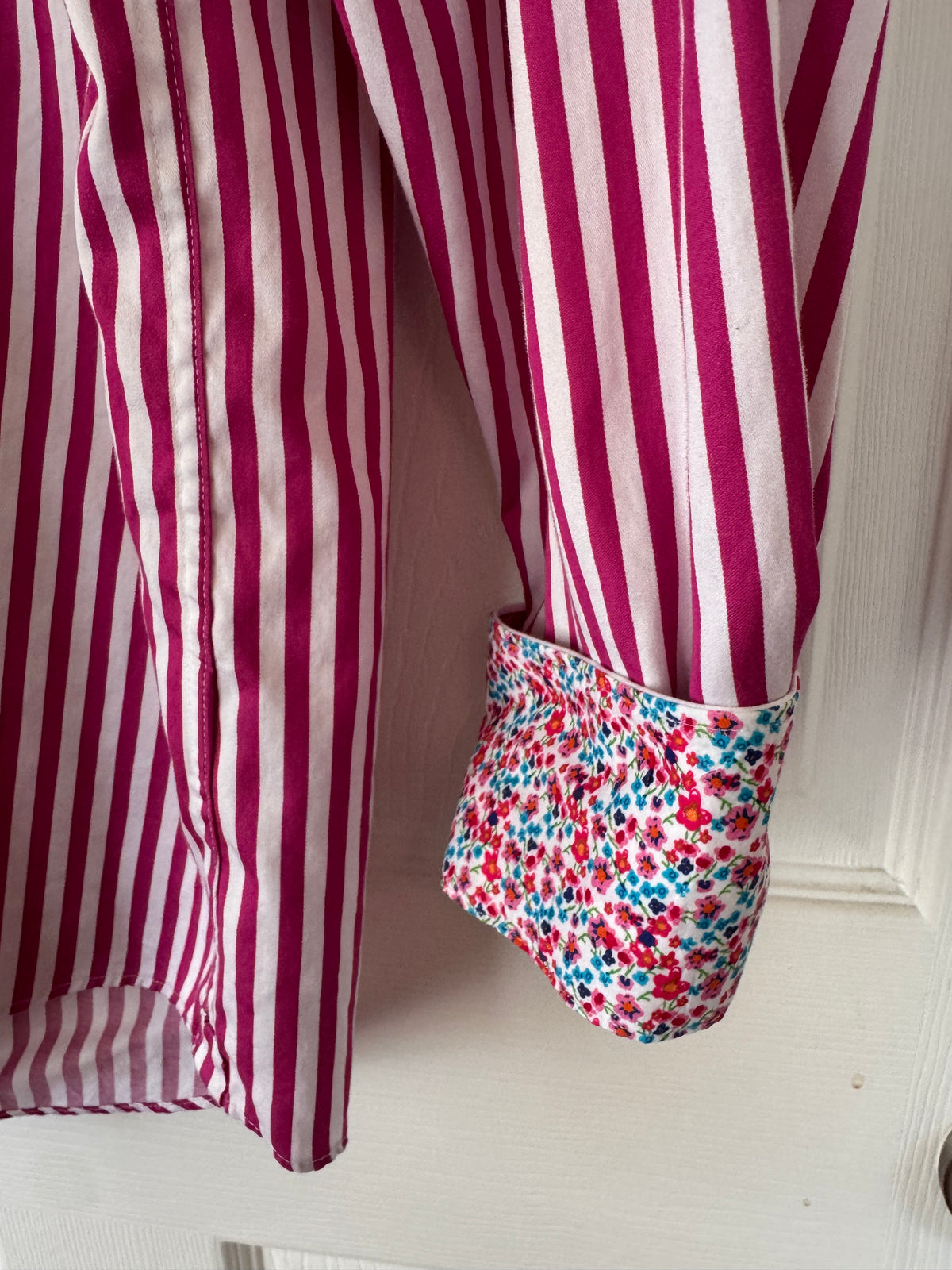 A close-up of a striped shirt with a rolled-up cuff showing a contrasting floral patterned lining, hanging against a white paneled door.