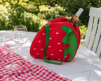 A red strawberry-shaped wine bottle cooler bag with green leaves and white polka dots sits on a white outdoor table next to two wine glasses and a red-and-white checkered cloth. A wine bottle is partially tucked inside.