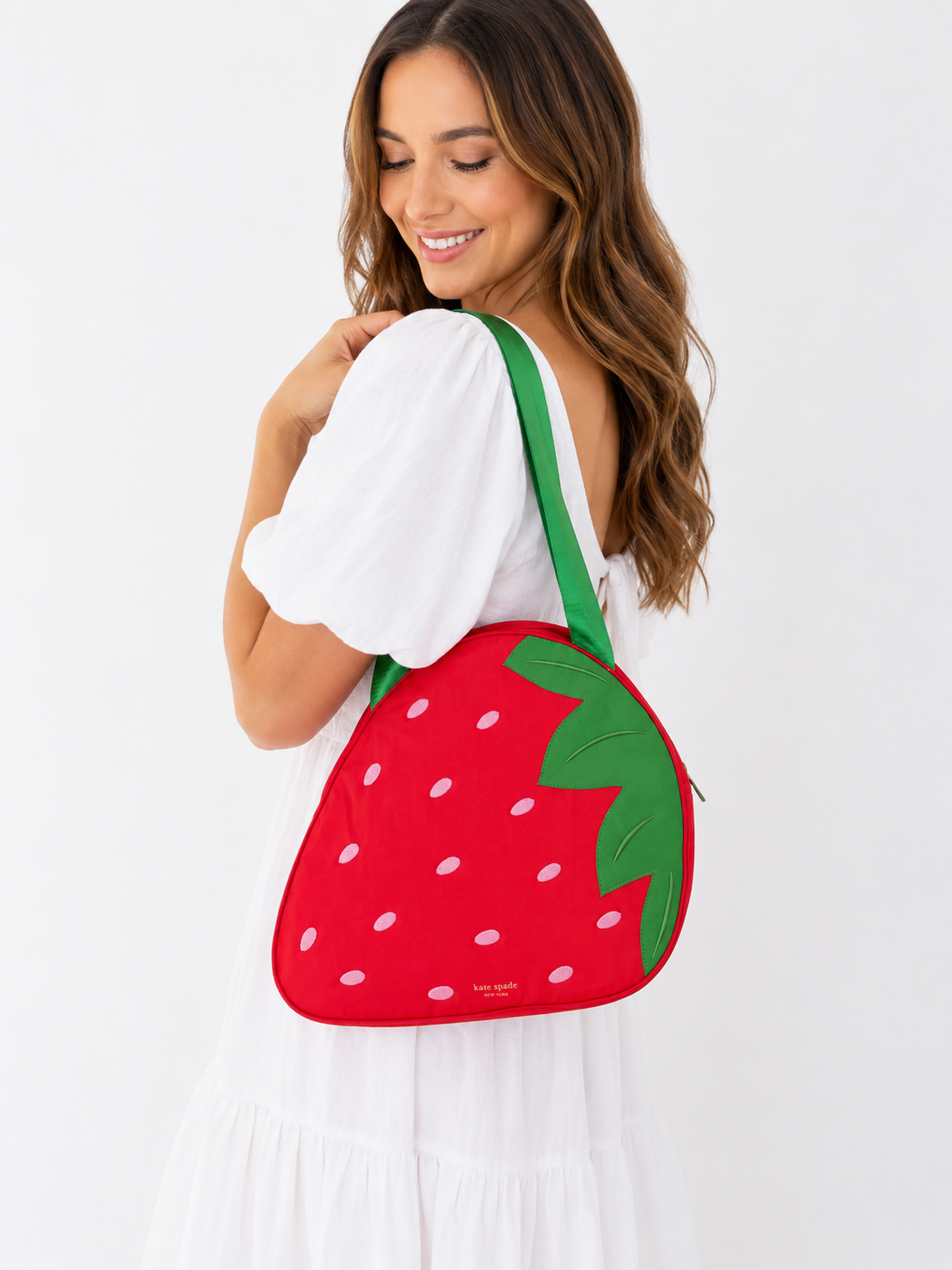A woman wearing a white dress carries a red strawberry-shaped shoulder bag with green straps and white embroidered details, set against a plain white background.