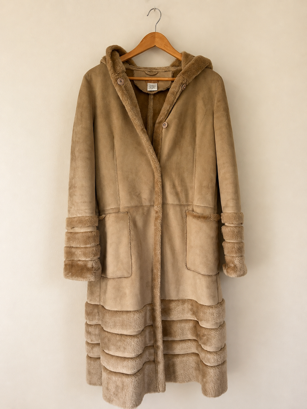 A mid-length hooded coat in tan suede-like material with contrasting brown faux-fur trim along the hood, cuffs, pockets, and hem, hanging on a wooden hanger against a plain beige wall.