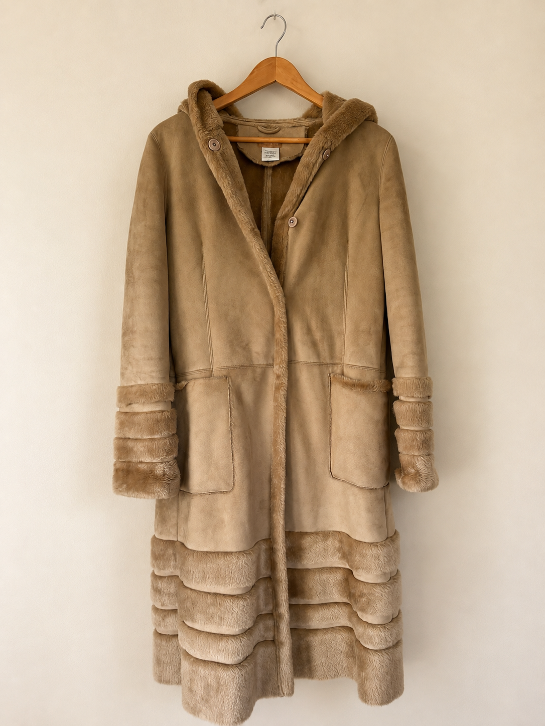 A mid-length hooded coat in tan suede-like material with contrasting brown faux-fur trim along the hood, cuffs, pockets, and hem, hanging on a wooden hanger against a plain beige wall.