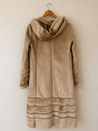 A beige faux fur coat with a large cowl neck and layered ruffle trim at the hem, hanging on a wooden hanger against a plain beige wall.